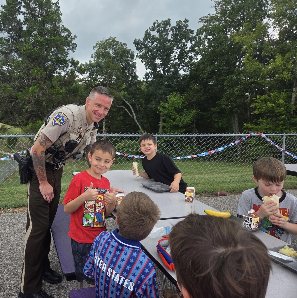 Uniformed law enforcement officer smiles beside elementary students eating lunch outdoors at picnic tables.