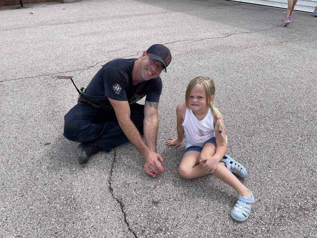 Firefighter and young student sit together outdoors, smiling at the camera during a school visit.