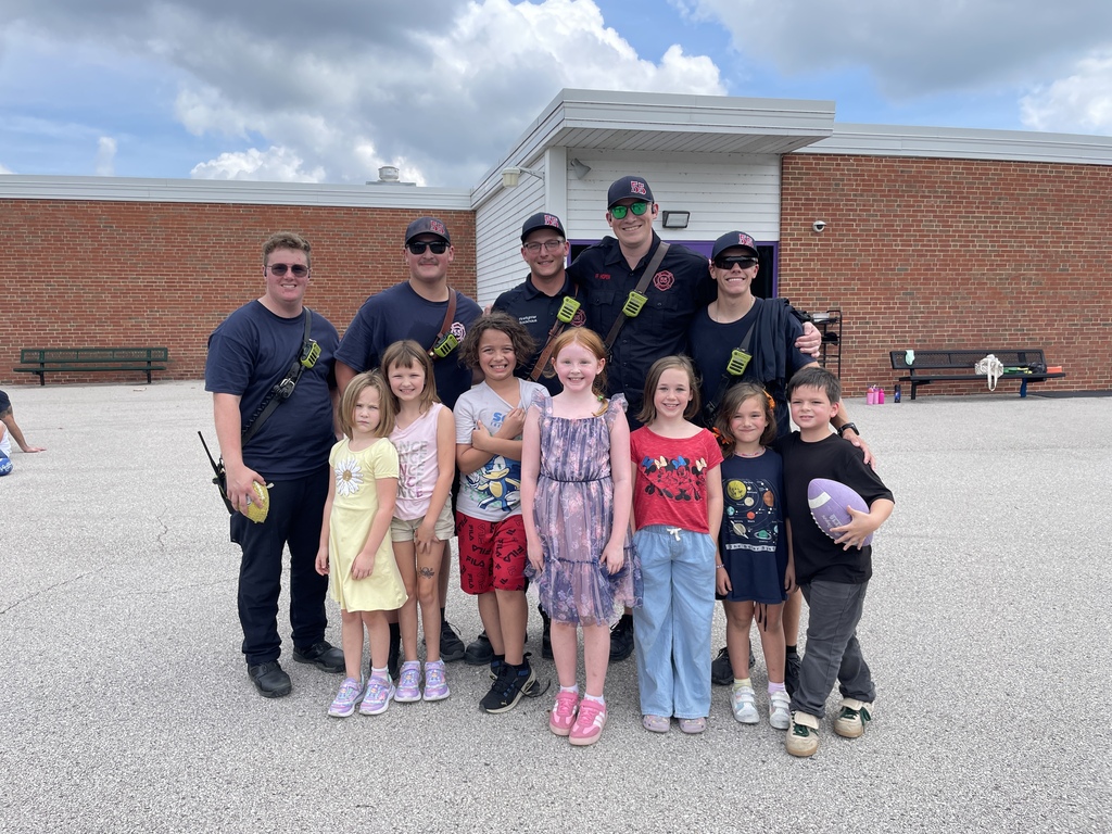 Five firefighters pose with seven smiling elementary students outside a brick school building.