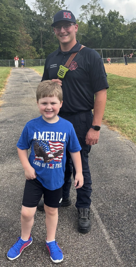 Firefighter poses outdoors with a smiling student in a blue patriotic shirt near a school playground.