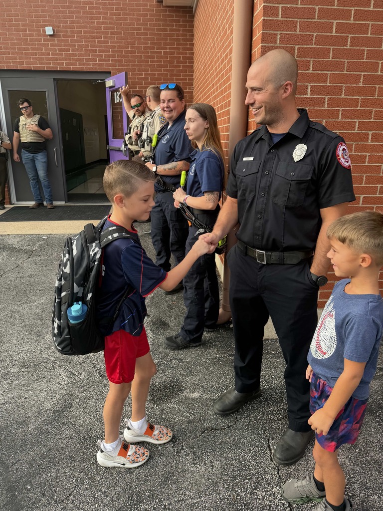 Student fist-bumps a smiling firefighter outside a school as first responders welcome arriving children.