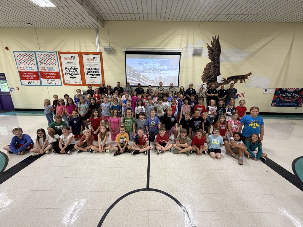 Large group of elementary students and first responders pose together beneath a Patriot Day display at school.