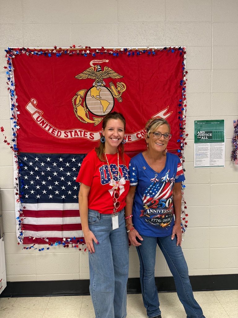Two school staff members in patriotic clothing smile in front of U.S. Marine Corps and American flag decorations.