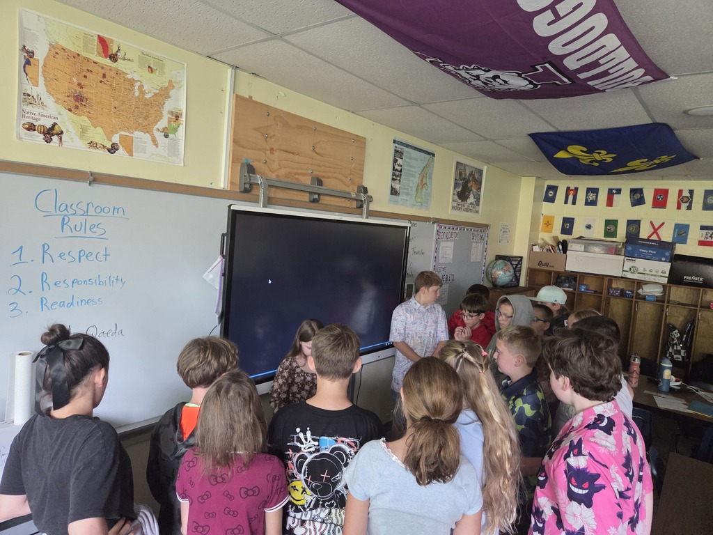 Students gather around a classroom display, with rules emphasizing respect, responsibility, and readiness on the whiteboard.