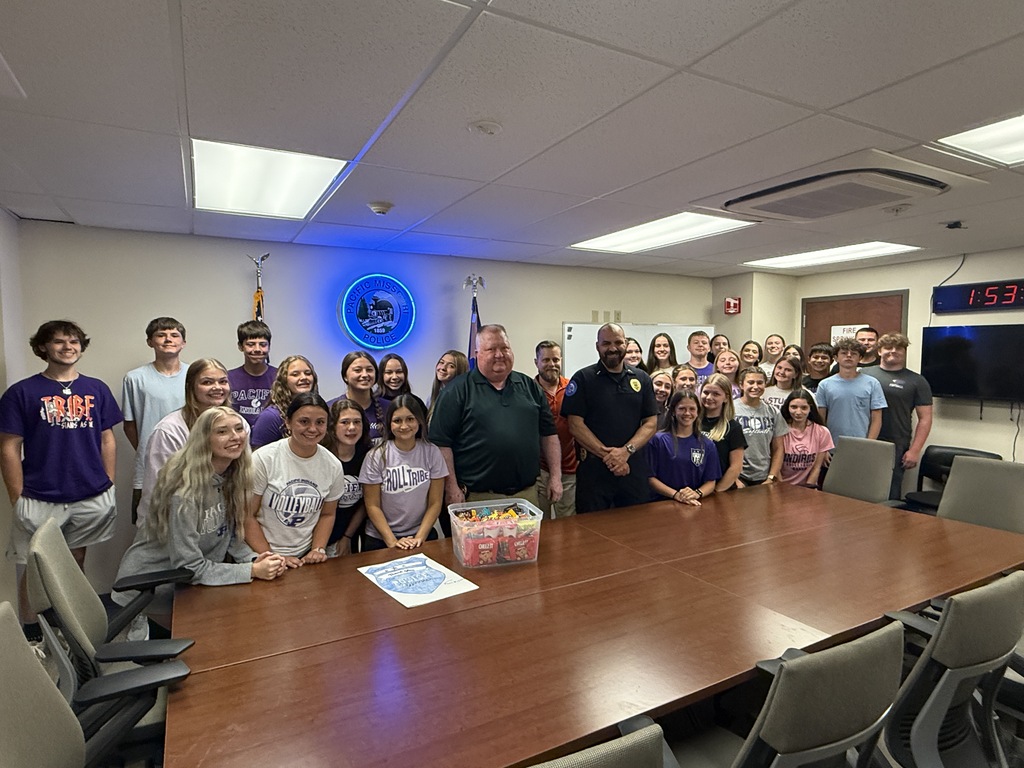 Students pose with police officers in a police department conference room after presenting a container of treats.