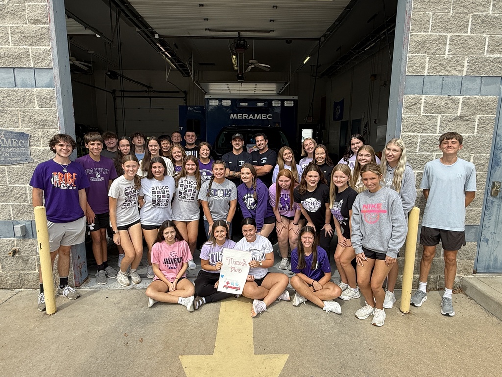 Students pose with first responders outside a fire station, with a handwritten thank-you sign displayed in front.