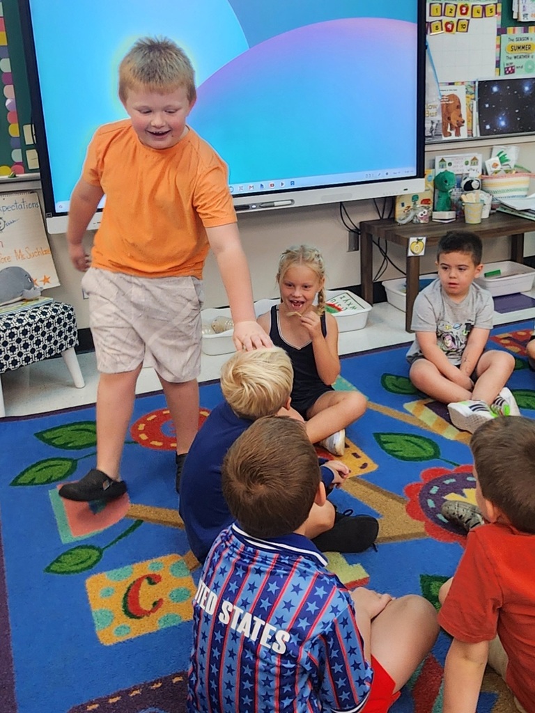 Student stands and reaches toward a seated classmate as other children watch during a classroom group activity.
