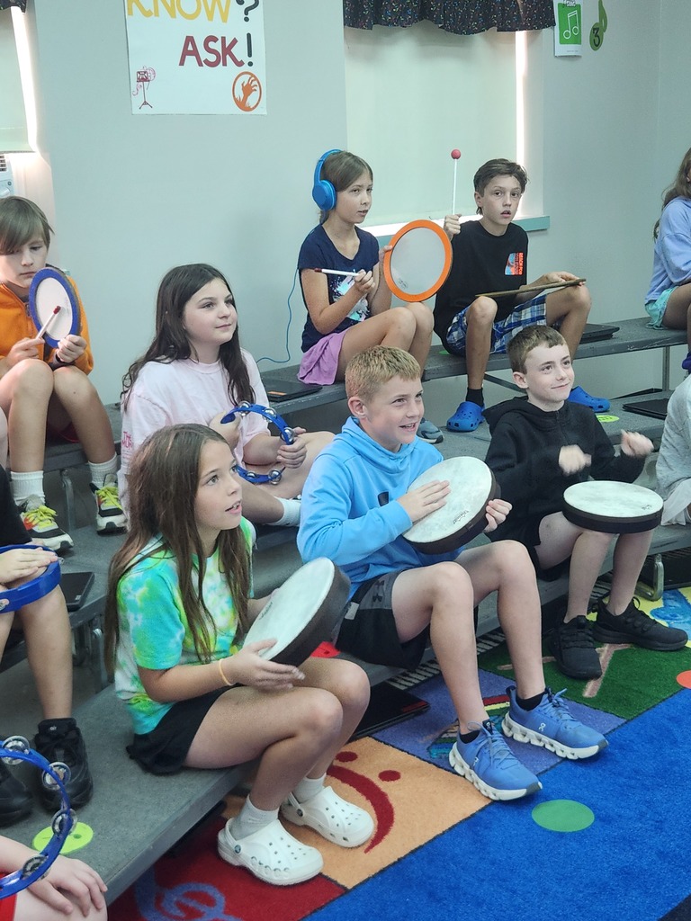 Students seated on classroom risers play hand drums, tambourines, and other percussion instruments together.