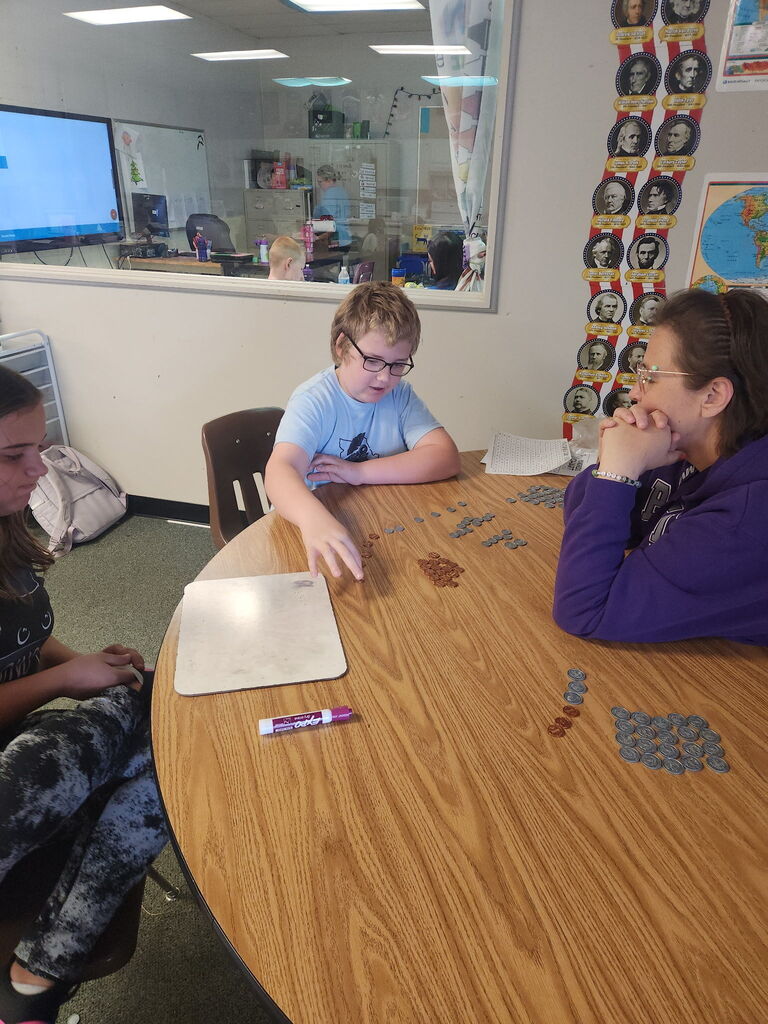 Students gather around a classroom table, sorting and counting groups of coins during a hands-on math activity.