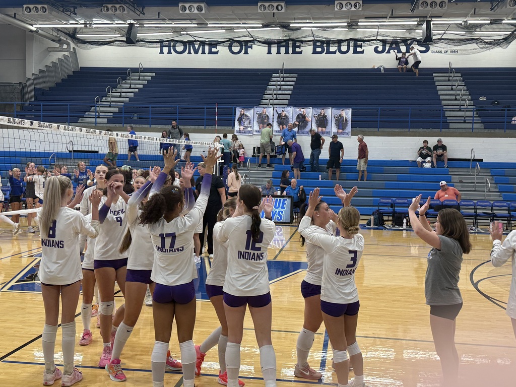 Volleyball players in white and purple uniforms exchange high-fives near the net during a match in a school gym.