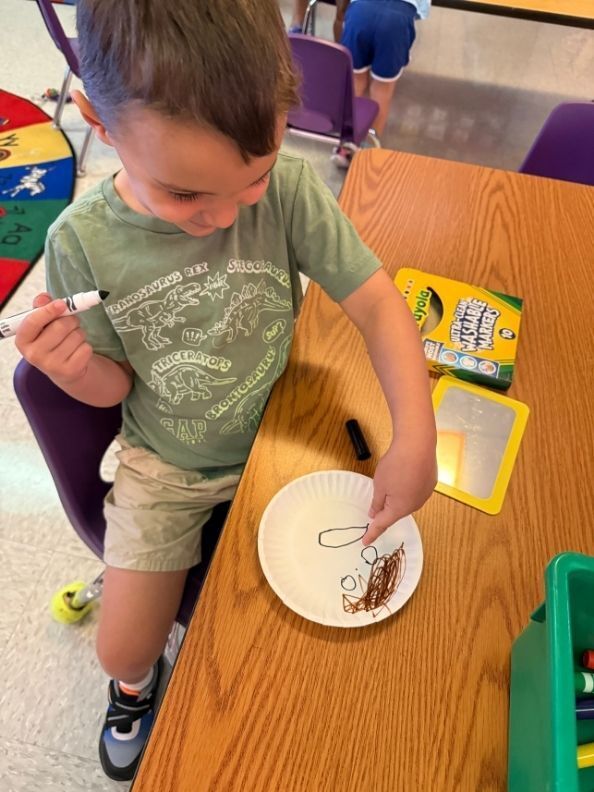 A student smiles while drawing on a paper plate with a marker during a classroom art activity.