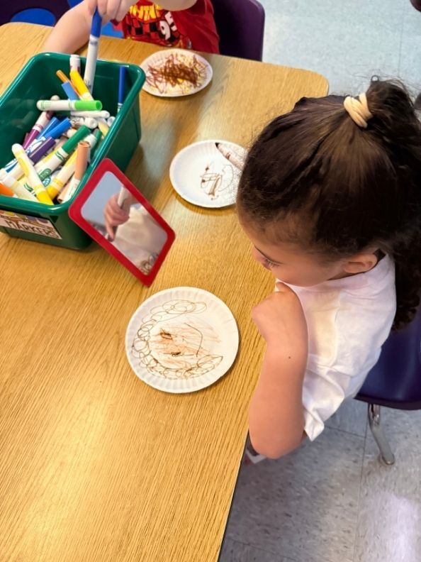 A student studies her reflection in a small mirror while drawing a self-portrait on a paper plate with markers.