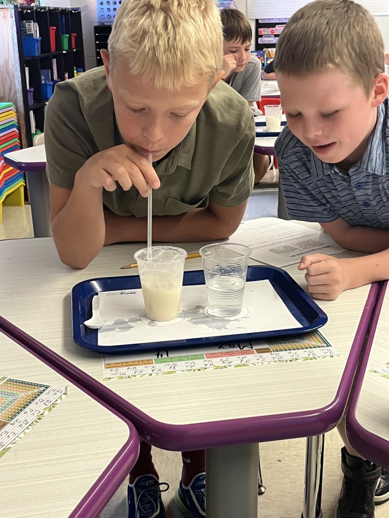 Two students observe cups of liquid as one blows through a straw during a classroom science experiment.