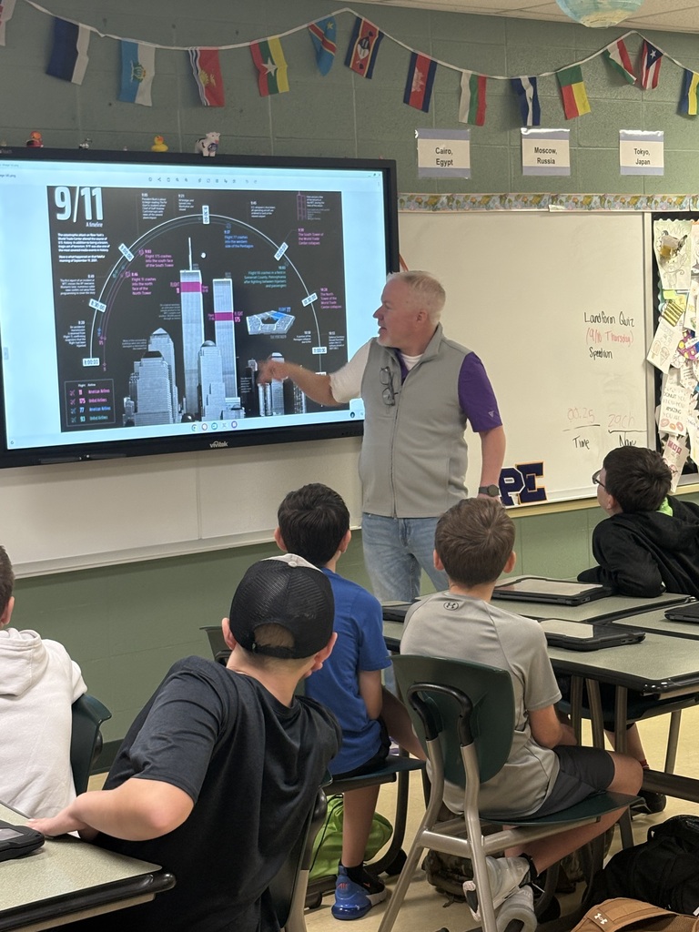 A teacher points to a 9/11 timeline displayed on a classroom screen as students watch from their desks.