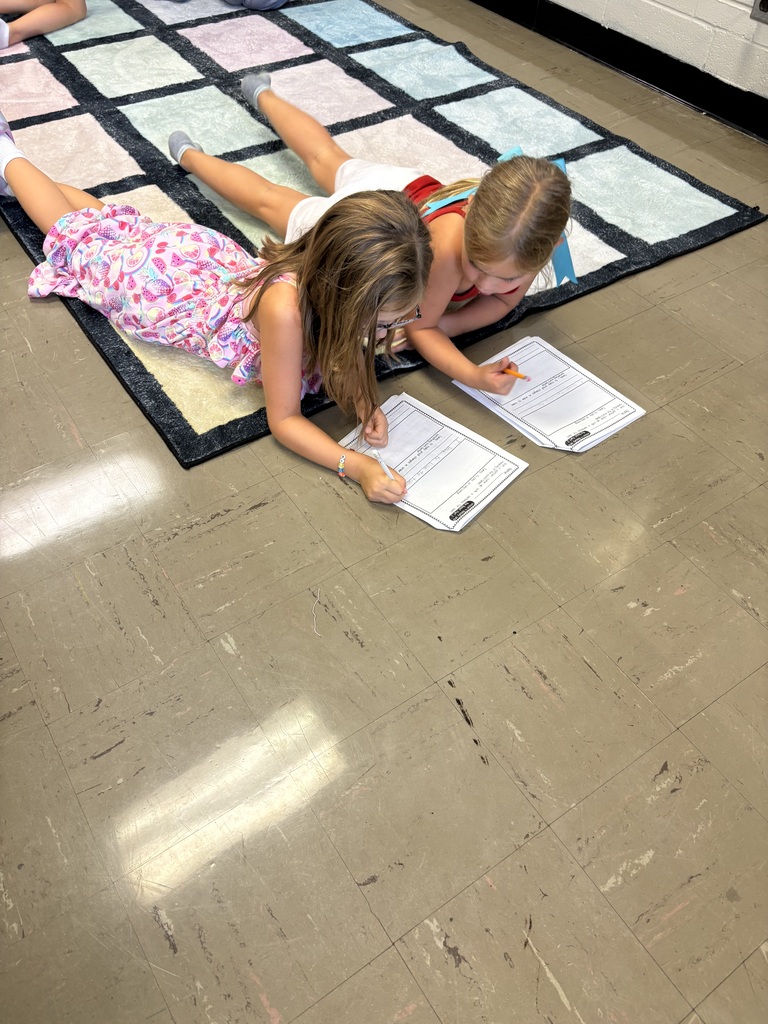 Two students lie on a classroom rug, writing on worksheets side by side with pencils.