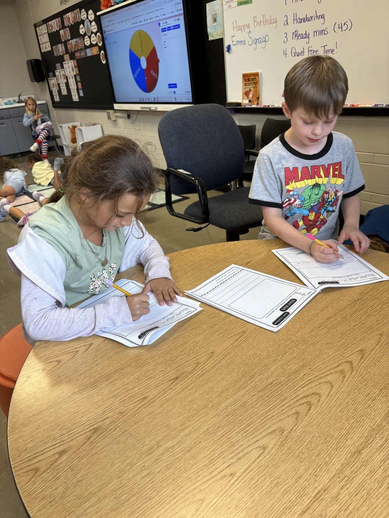Two students sit at a classroom table, concentrating as they complete handwriting worksheets.