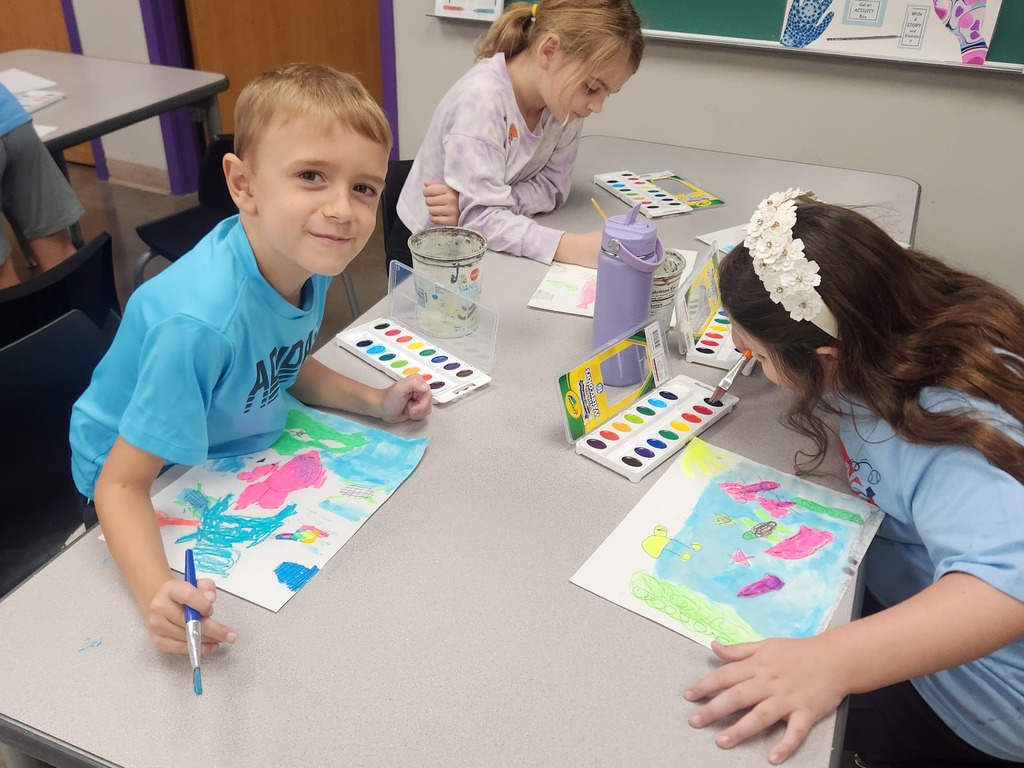 Three students paint colorful underwater artwork with watercolor sets at a classroom table.