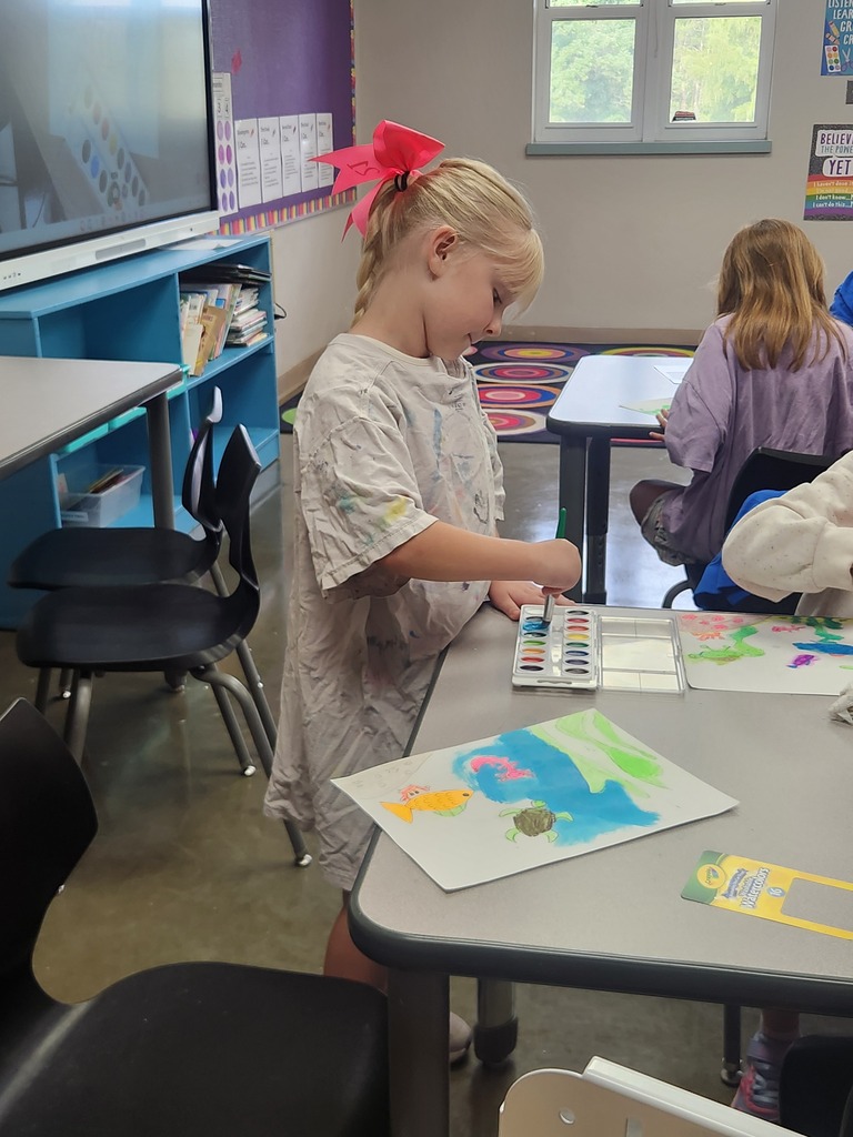 A student with a bright pink bow paints an underwater scene with fish and a turtle using watercolors.
