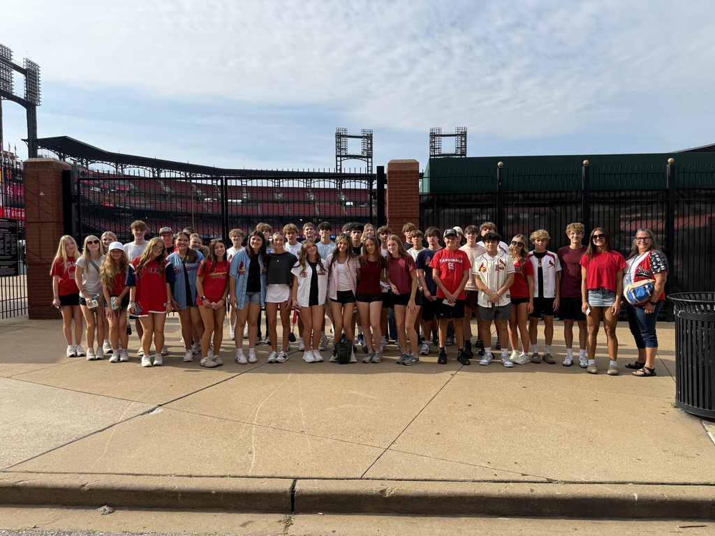 A large group of students and adults pose together outside a baseball stadium, many wearing red team apparel.