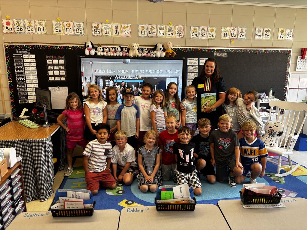 A teacher and a class of smiling students pose together after a classroom read-aloud activity.