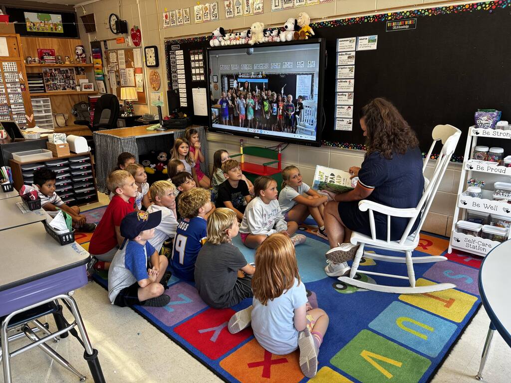 A teacher reads a picture book aloud while students sit attentively together on a colorful classroom rug.