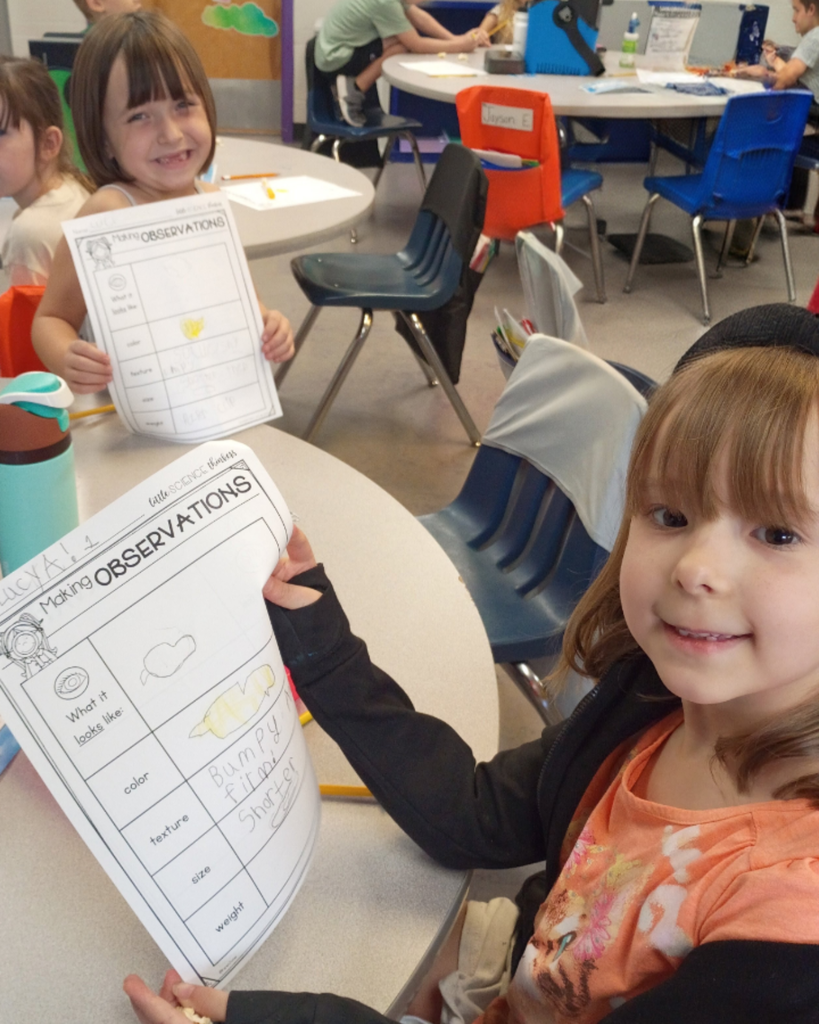 Two smiling elementary students hold up their completed science observation worksheets while sitting at classroom tables.