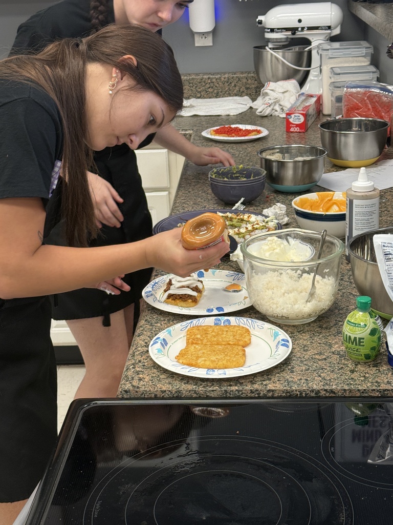 Two students prepare a meal in a kitchen, adding toppings to crispy hash browns surrounded by bowls of ingredients.