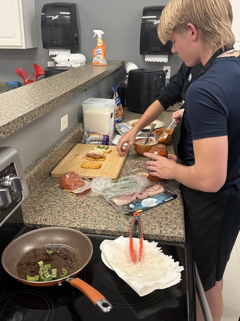 A student in a black apron assembles a meal with sauce, cheese, and vegetables at a kitchen counter.