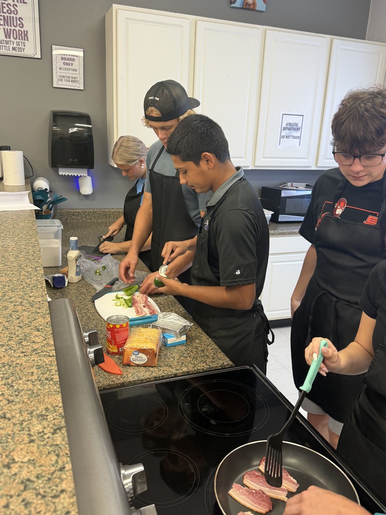 Four students work together in a kitchen, preparing ingredients and cooking bacon on the stovetop.