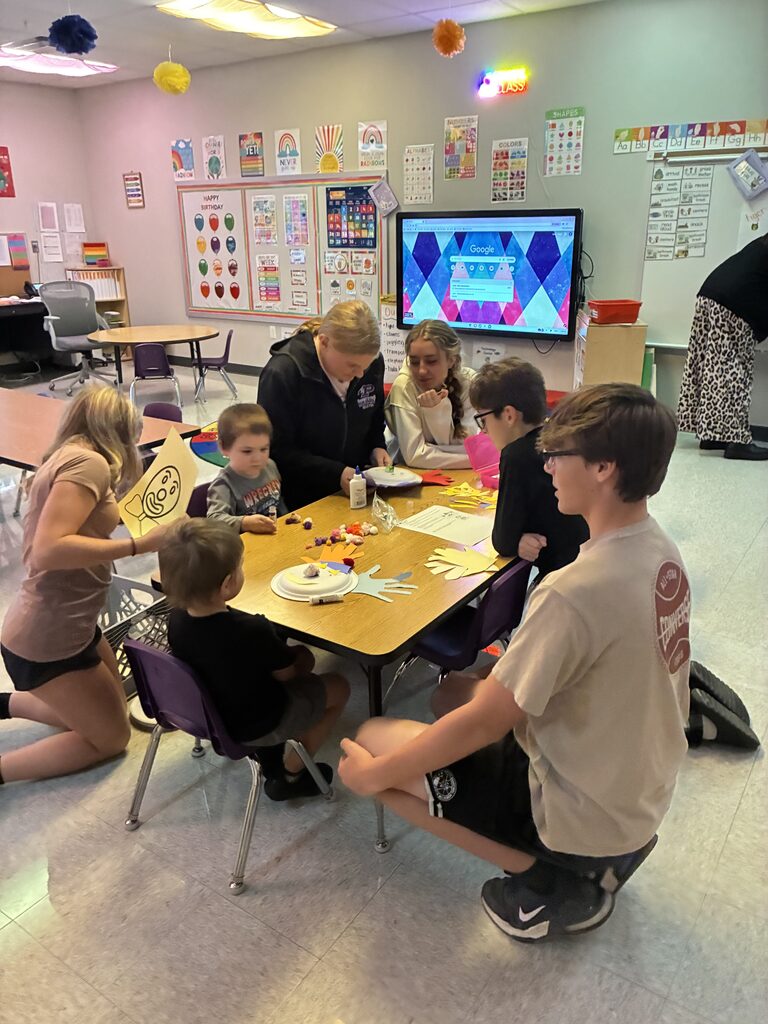Older students help younger children create colorful paper crafts and decorations around a classroom table.