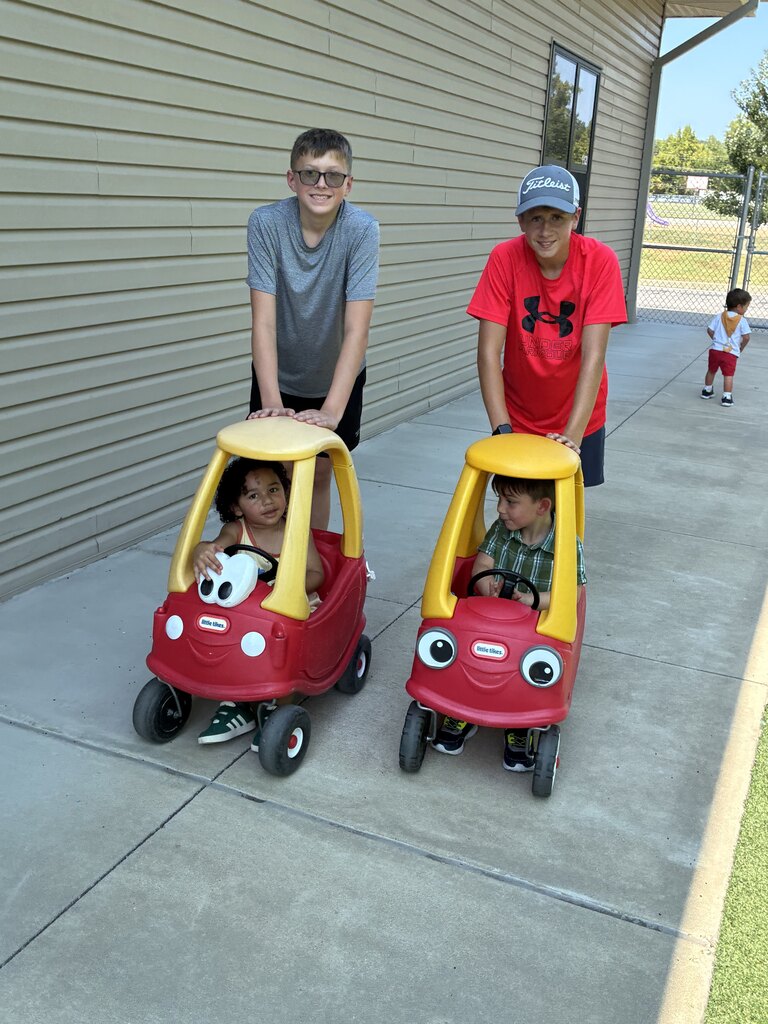Two smiling teens push younger children in red and yellow toy cars along a sunny outdoor walkway.