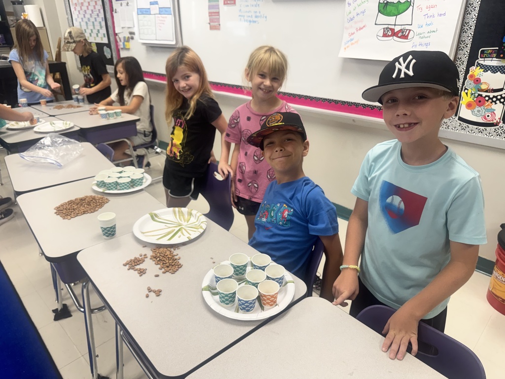 Smiling elementary students gather around classroom tables, sorting dried beans into colorful paper cups for a hands-on activity.