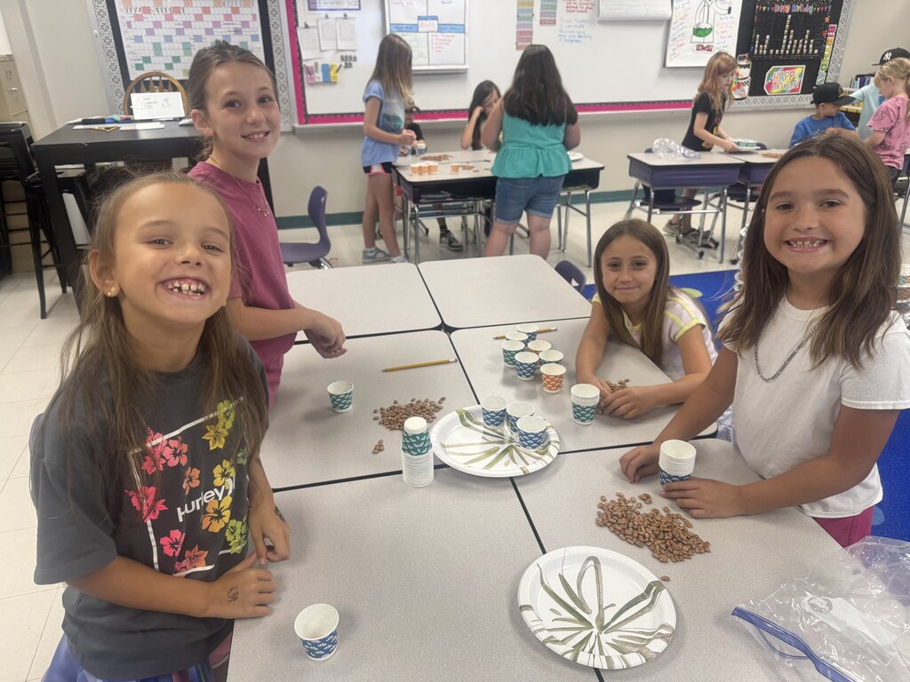 Four smiling students stand around a classroom table with dried beans, paper cups, and plates during a group activity.
