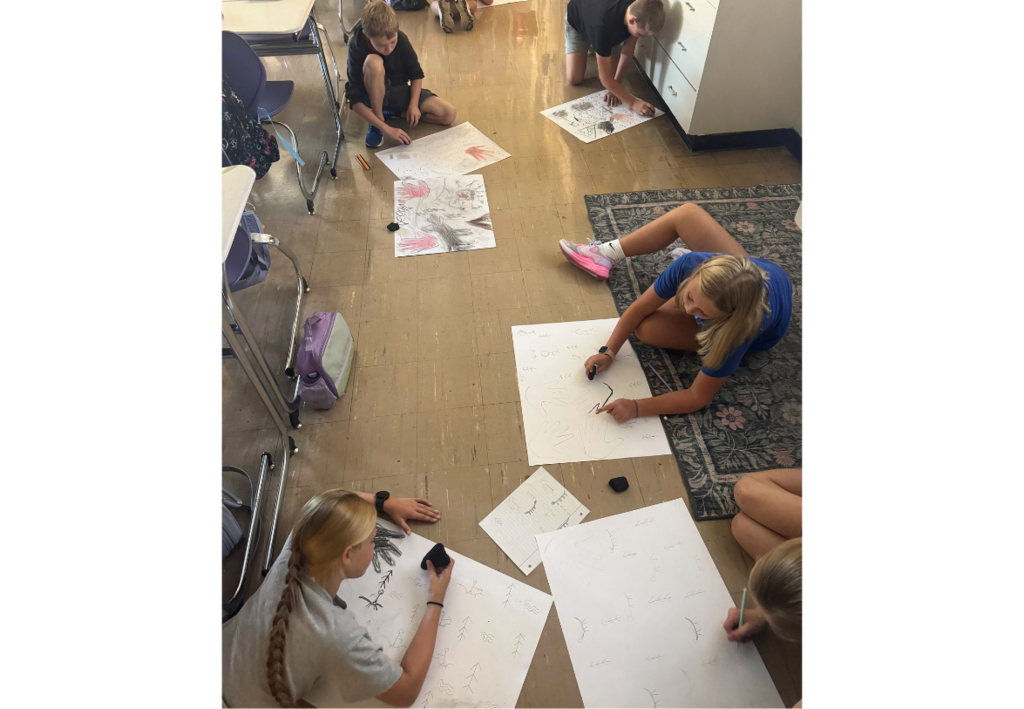 Students sit on a classroom floor, working together on large paper drawings with pencils and markers.