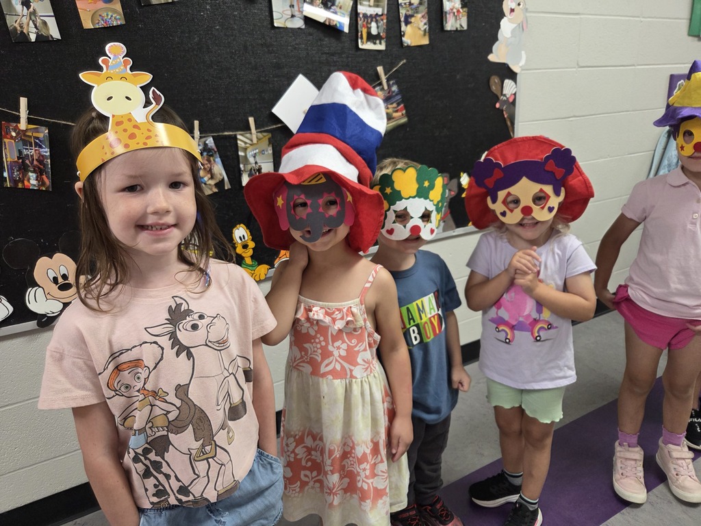 Preschoolers smile and pose in colorful animal and clown masks and playful hats in a school hallway.
