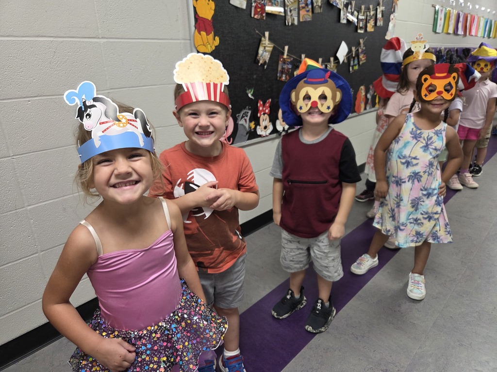 Smiling preschoolers show off colorful circus-themed hats and masks while standing together in a school hallway.