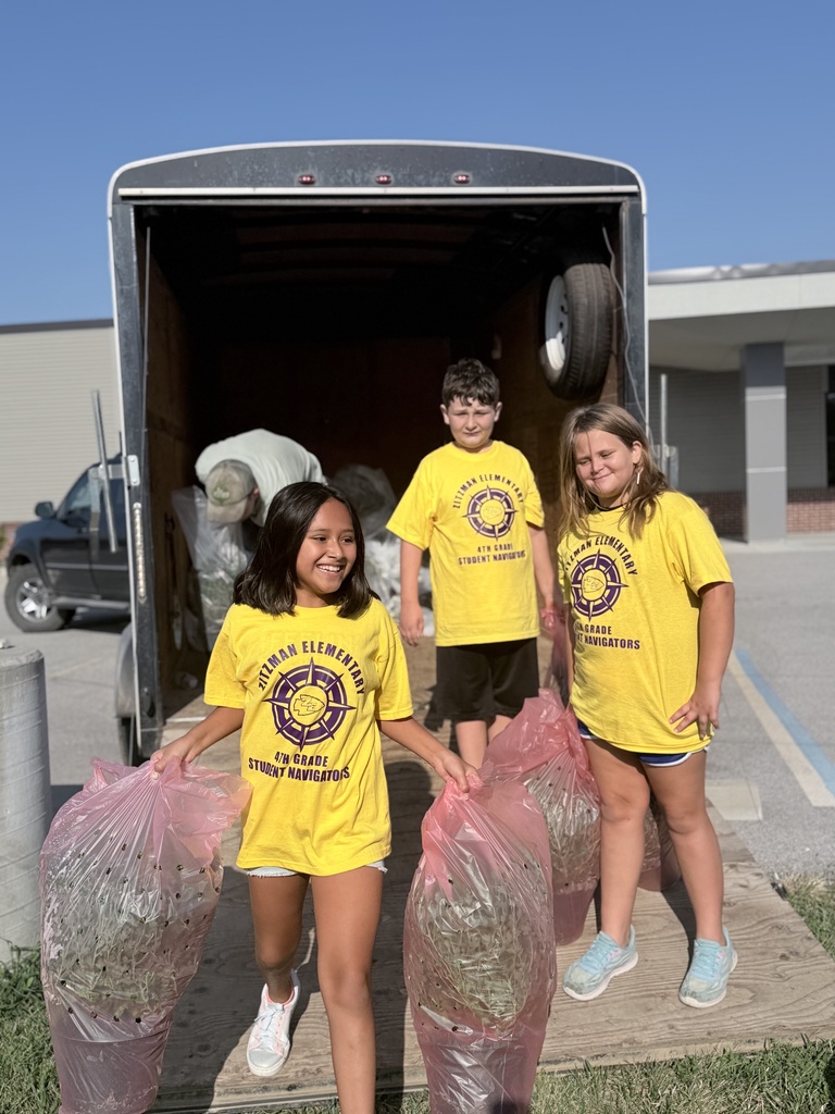 Three elementary students in yellow Student Navigator shirts carry large bags of yard waste from a trailer during a cleanup project.