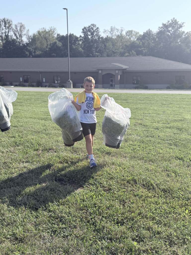 A smiling student carries two large bags of collected grass clippings across a sunny field during an outdoor cleanup.