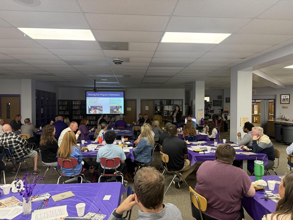 Audience members seated at purple-covered tables watch a presenter discuss program celebrations in the school library.