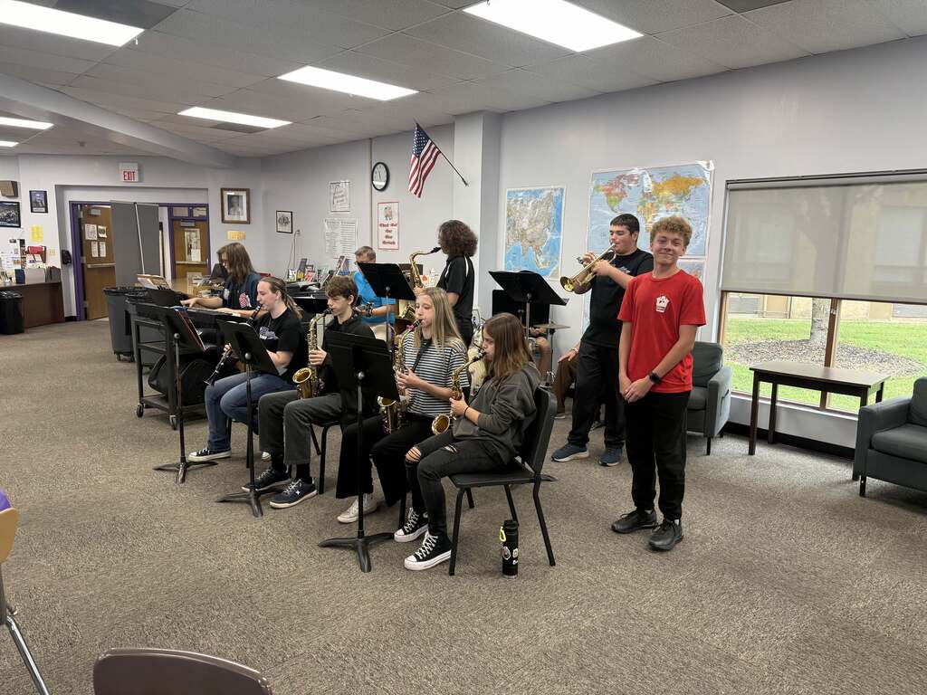 Student musicians perform on saxophones, clarinet, trumpet, and keyboard during an event in the school library.