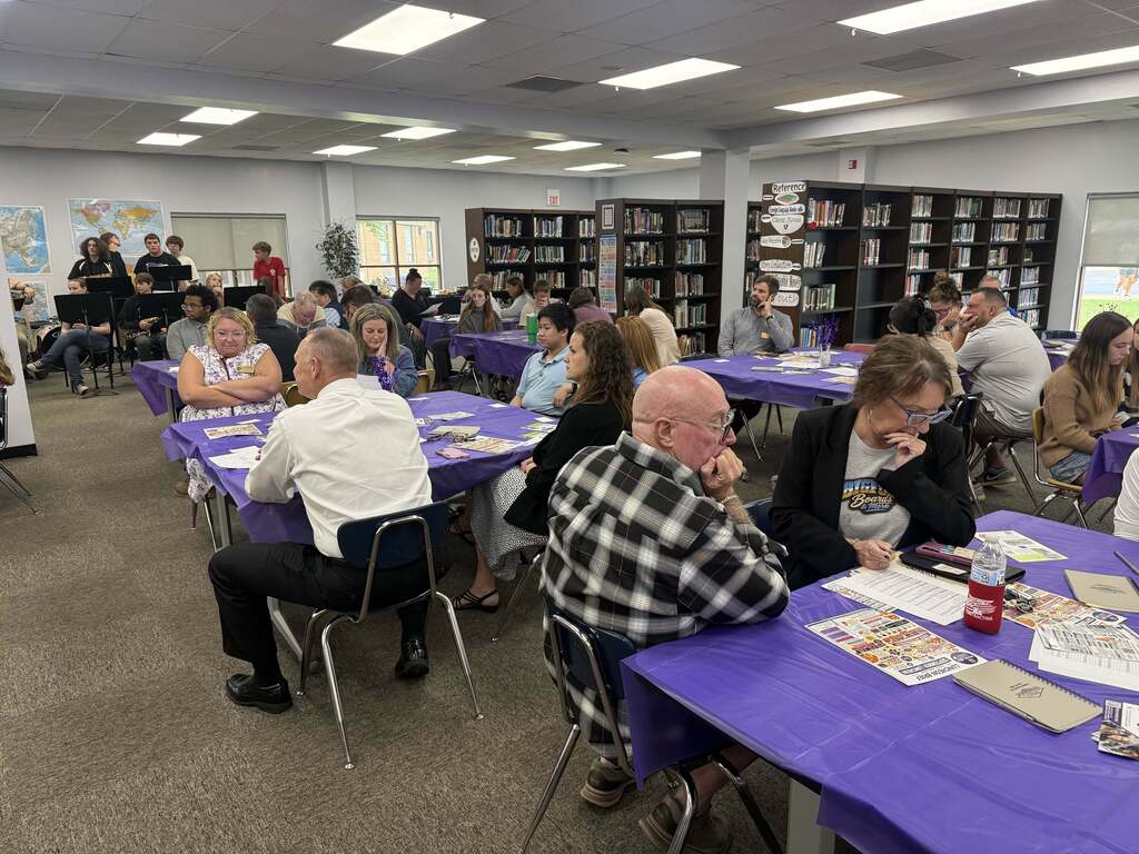 Adults and students gather at purple-covered tables in a school library while a student music ensemble performs nearby.