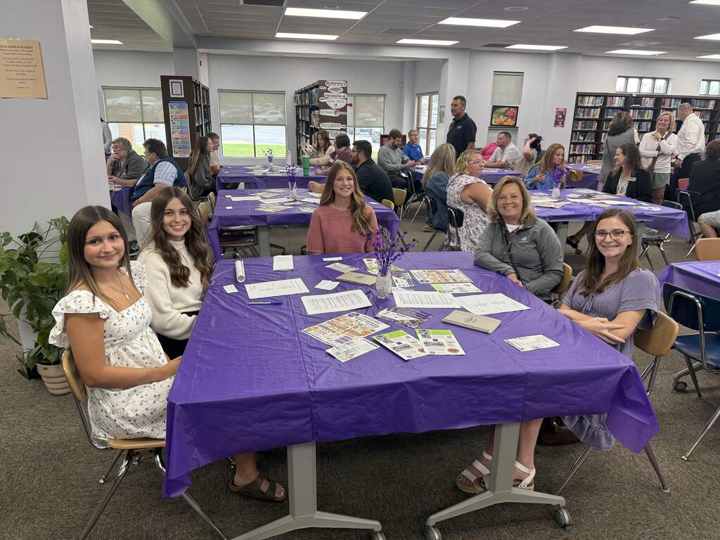 Five smiling students and adults pose around a purple-covered table during a gathering in the school library.