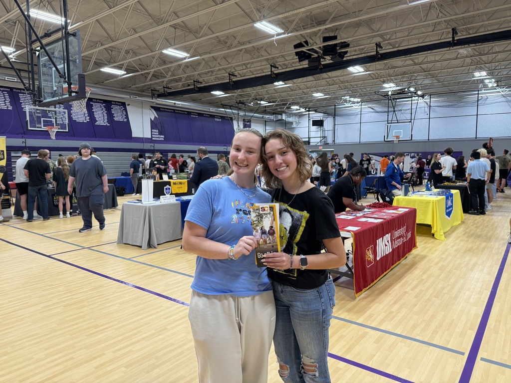 Two smiling students pose with a college brochure at a school college fair held in a gymnasium.