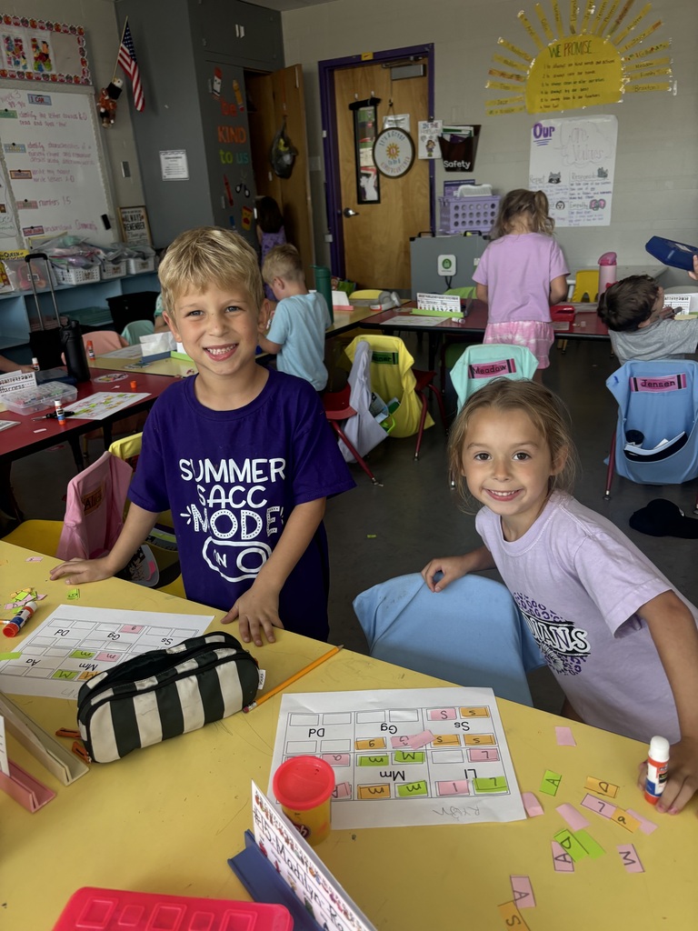 Two smiling students stand beside a classroom table covered with letter-matching worksheets and colorful paper pieces.