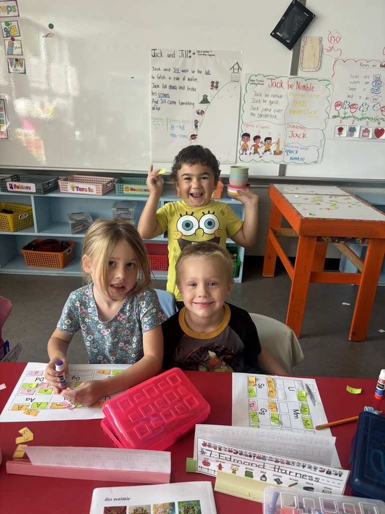 Three smiling students work on a colorful letter-matching activity at a classroom table while another student holds tape rolls.