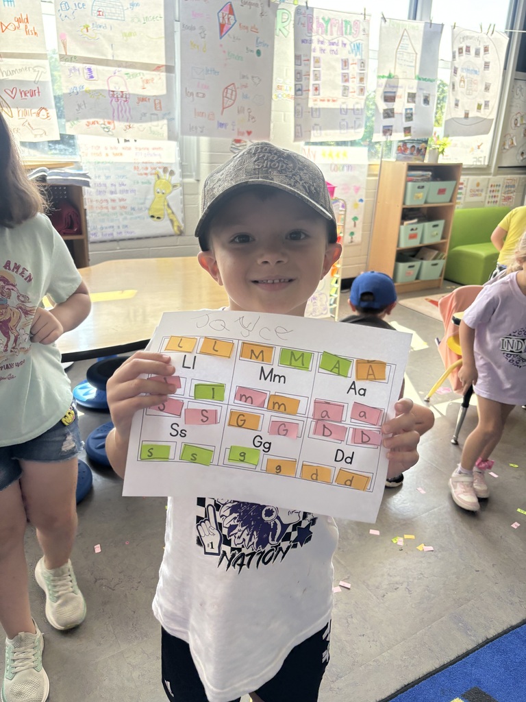 A smiling student wearing a baseball cap proudly holds up a completed letter-matching worksheet in a bright classroom.