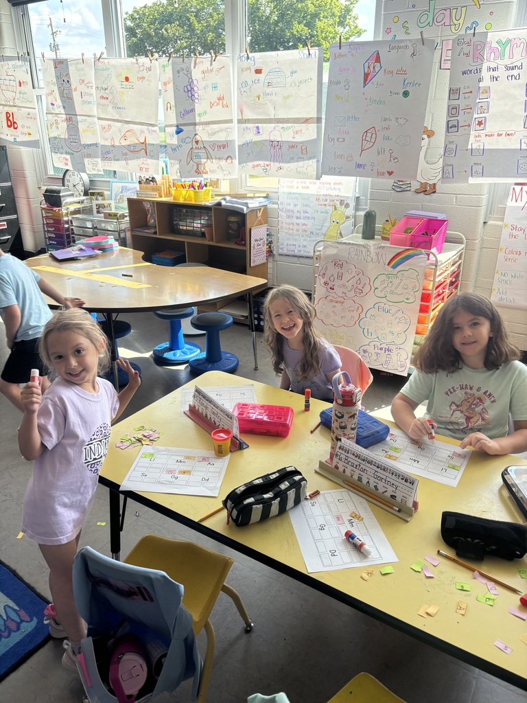 Three students smile around a classroom table while completing hands-on letter-matching activities with colorful paper tiles.