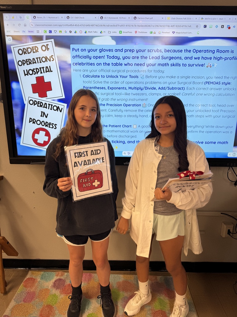 Two students pose with first-aid and patient materials in front of an “Order of Operations Hospital” classroom display.