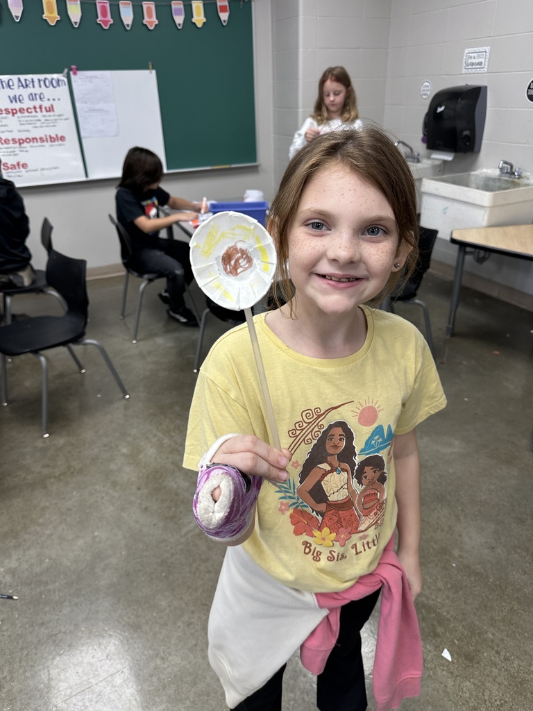 Smiling student in an art classroom holds up a handmade circular artwork mounted on a wooden stick.