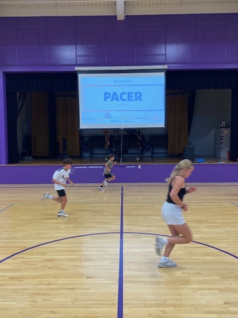 Students run across a school gym floor during the PACER fitness test as a timer and PACER screen are projected onstage.